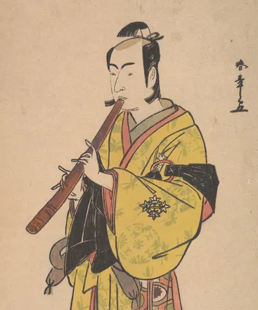 An actor dressed as a wandering komusō monk stands playing a long bamboo shakuhachi, his basket hat at his feet.